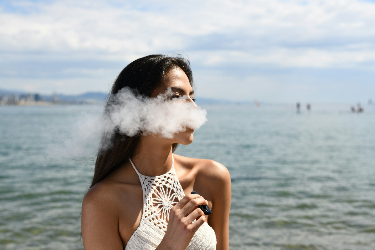 A woman exhales a cloud of vapor while holding a vape, similar to the AtingBar DF70 Disposable Vape, in her hand and a large expanse of water in the background