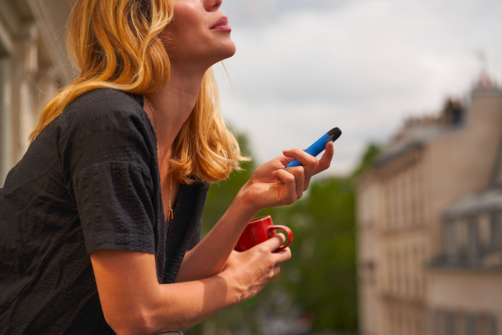 Woman holding a vape pen and a red cup, looking thoughtful while sitting on a balcony.